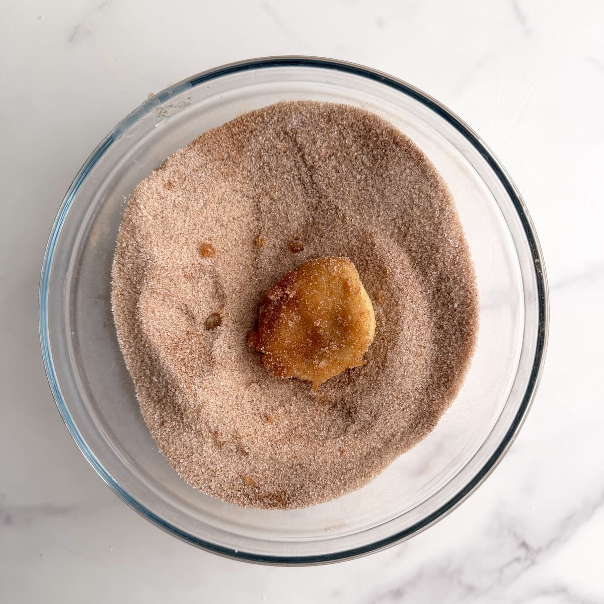A piece of sourdough monkey bread dough being coated in a bowl of cinnamon sugar with a marbled background.