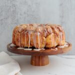 Sourdough monkey bread with icing dripping off the top featured on a beautiful wooden cake stand. The cake stand is on a white marble counter with a dark marble background. A white cloth is on the counter next to the cake stand.