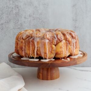 Sourdough monkey bread with icing dripping off the top featured on a beautiful wooden cake stand. The cake stand is on a white marble counter with a dark marble background. A white cloth is on the counter next to the cake stand.