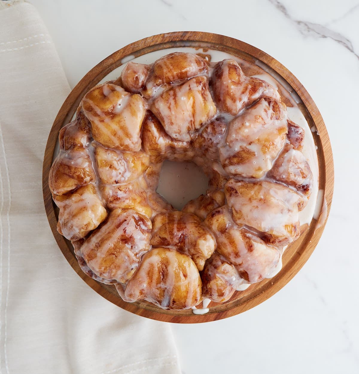 A top-down photo of the monkey bread displayed on a wooden cake stand. The cake stand is on a white marble counter with a white cloth laid diagonally next to the stand.