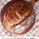 Loaf of cinnamon honey butter bread on a round wooden board with cinnamon sticks around it and next to a bowl of cinnamon honey butter.