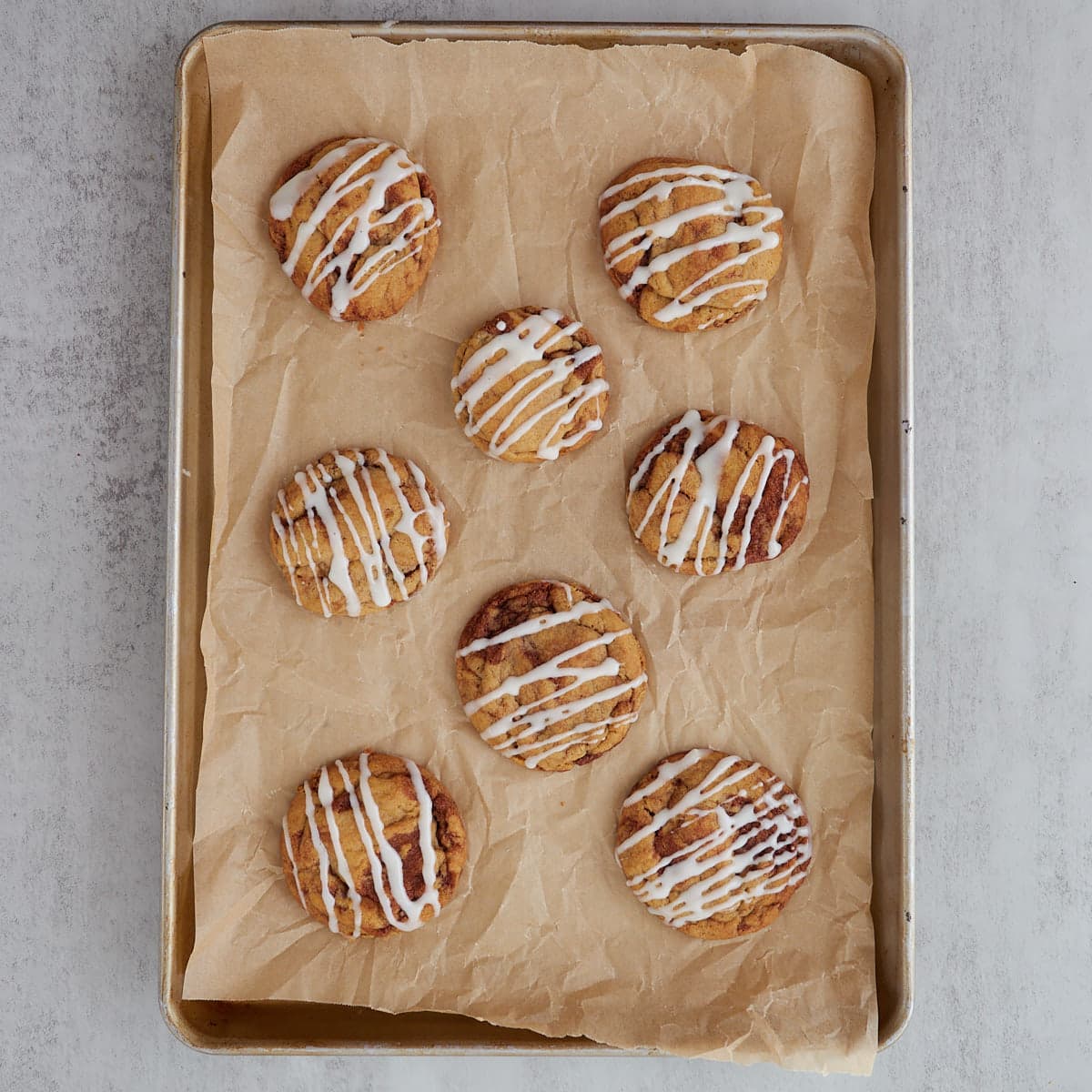 Eight sourdough cinnamon roll cookies on a parchment lined baking sheet after baking with cream cheese icing drizzled on top.