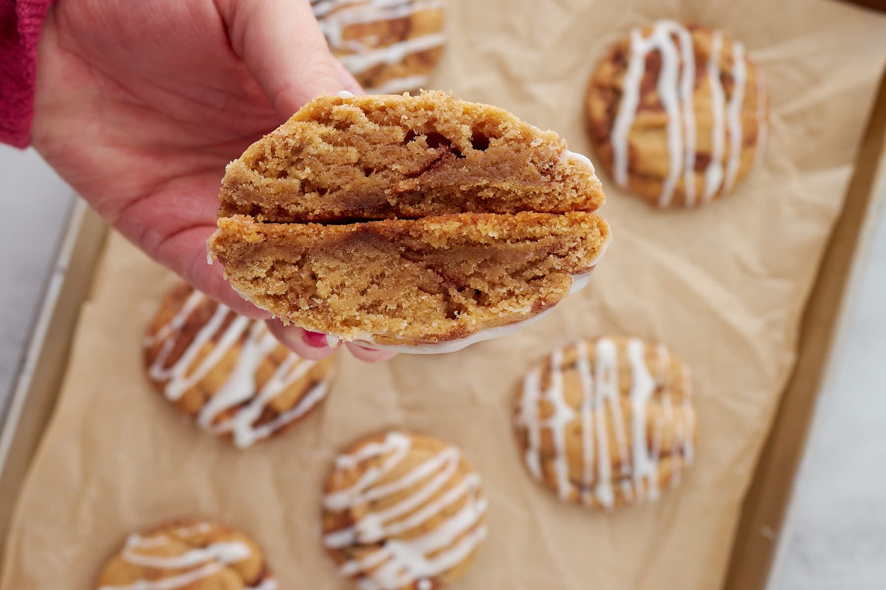 A close up view of a hand holding a sourdough cinnamon roll cookie broken in half. A parchment lined tray of cookies is behind the hand.