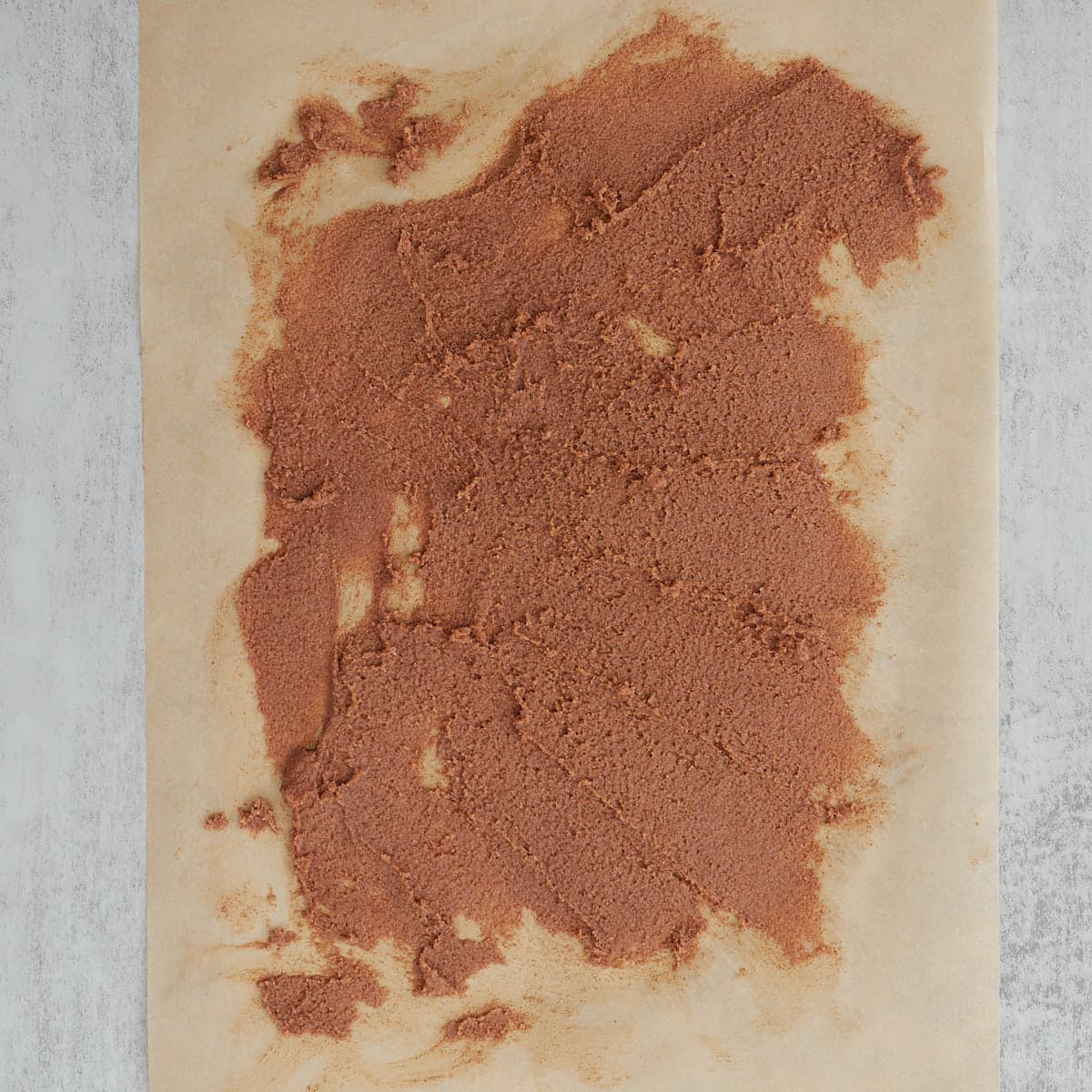 A thin layer of the brown sugar, butter, and cinnamon mixture for sourdough cinnamon roll cookies spread on a piece of parchment paper.