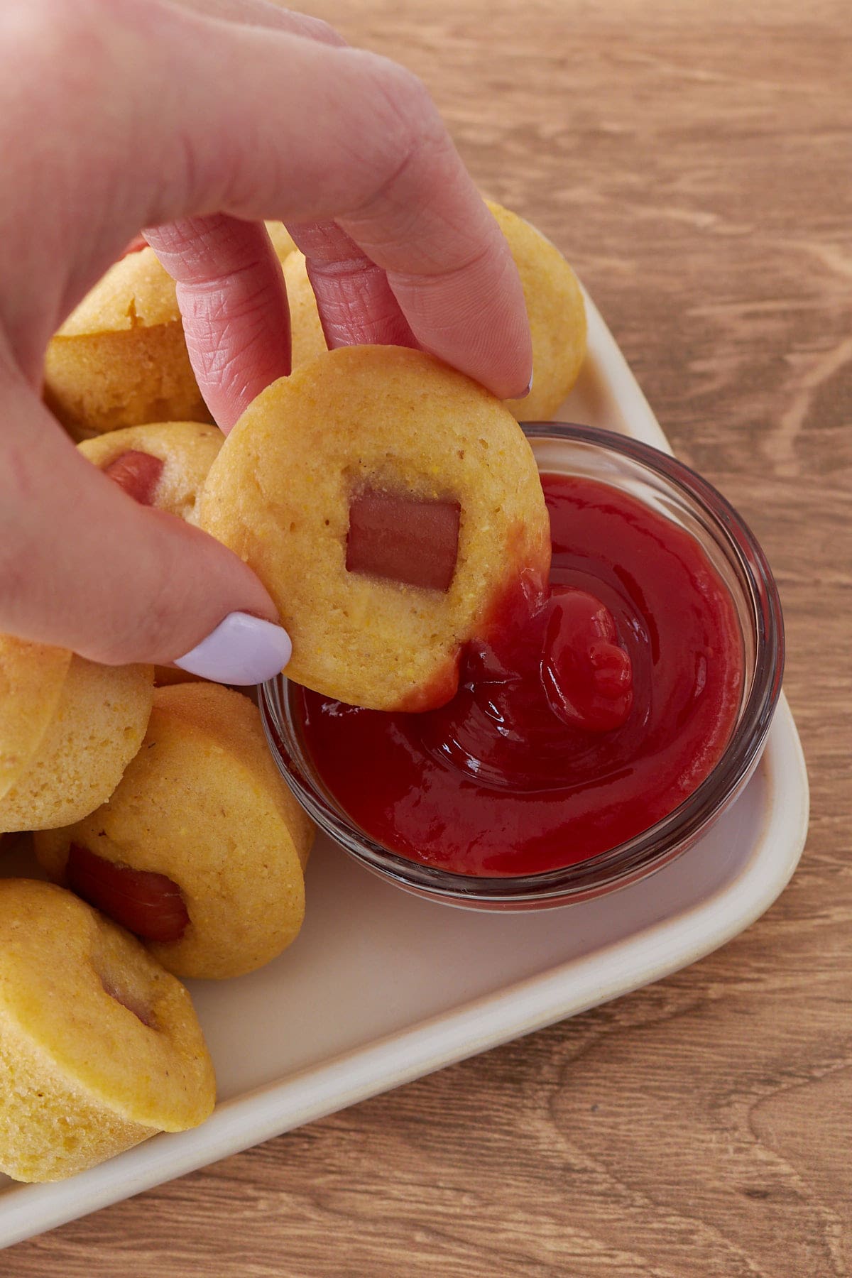 Hand dipping a Sourdough Mini Corn Dog into a small bowl of ketchup.