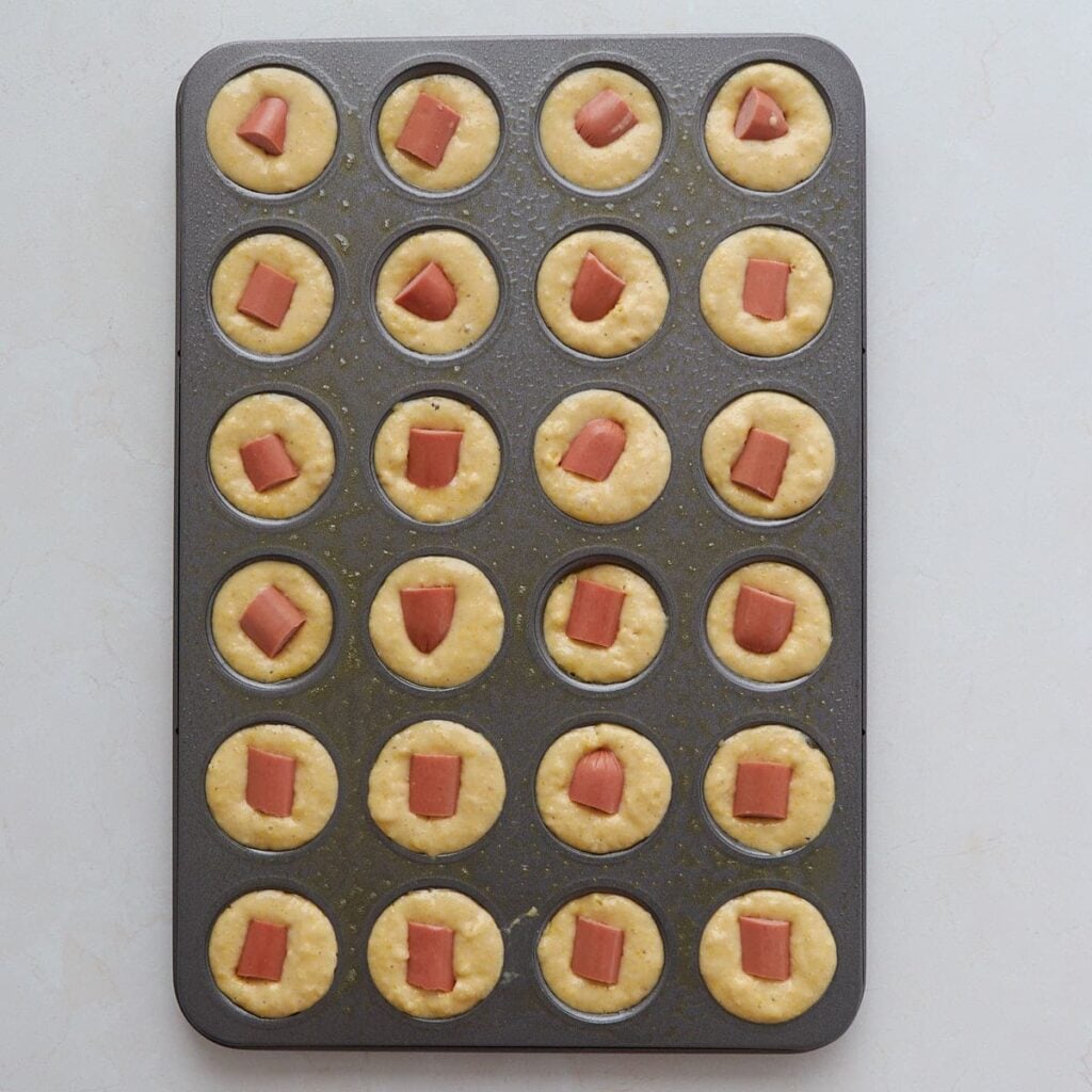 Hot dogs added to the corn bread batter in a mini muffin tin.