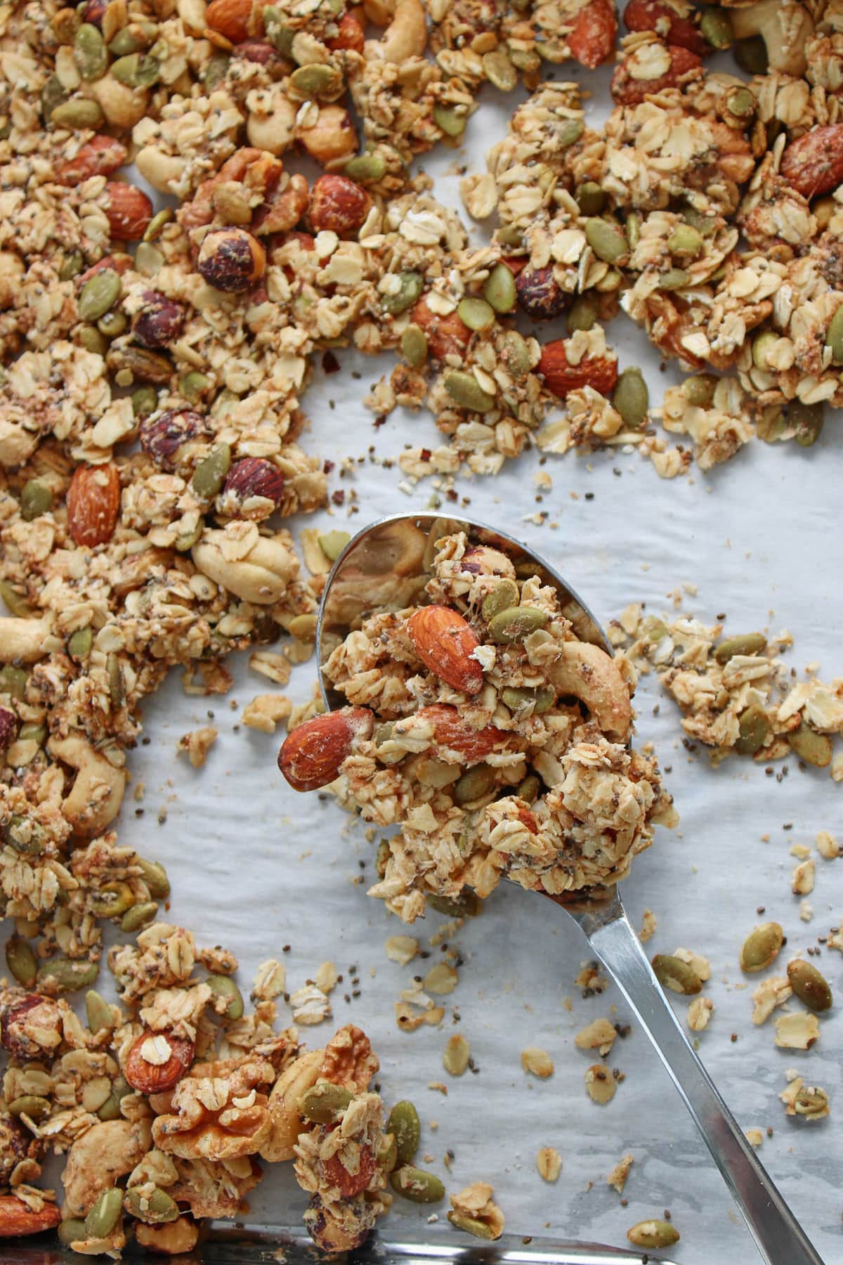 Silver spoon holding sourdough granola on a sheet pan lined with parchment paper filled with granola.