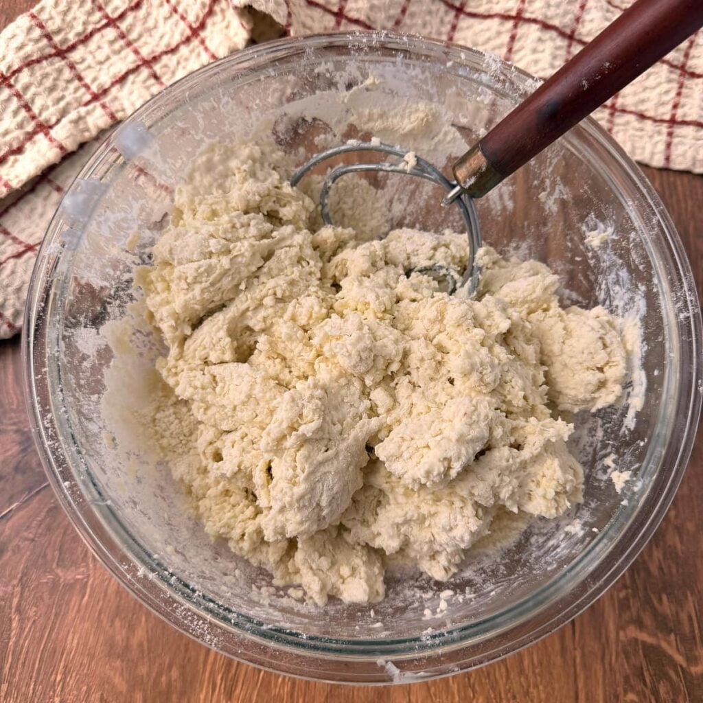 Dough for Sourdough Buttermilk Biscuits mixed in a clear bowl with a dough whisk.