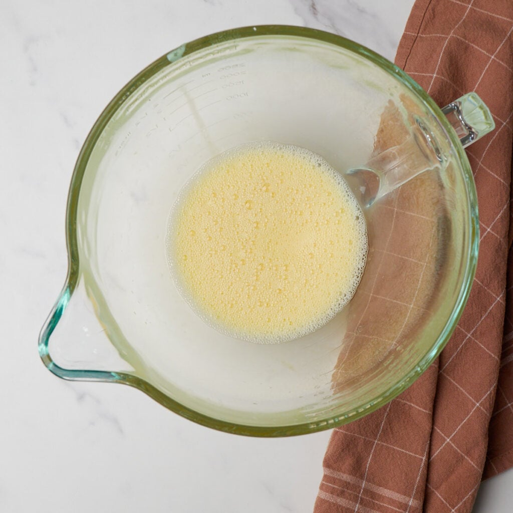 Eggs whisked in a clear bowl next to a brown towel.