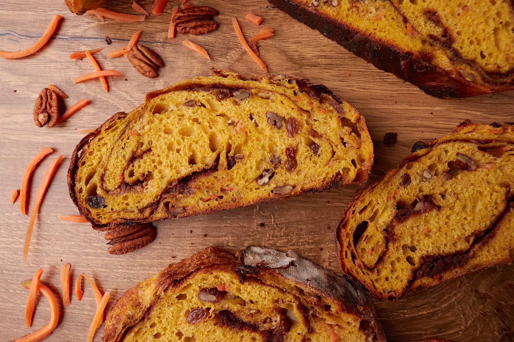 Slices of Carrot Cake Sourdough Bread on a wooden surface with shredded carrots and pecans scattered around.