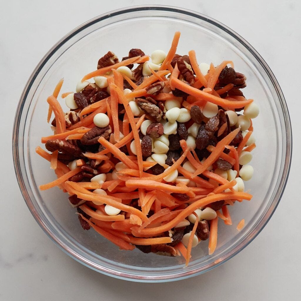Shredded carrots, pecans, raisins and cream cheese chips mixed together in a clear bowl.