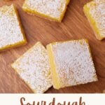 Squares of sourdough lemon bars on a wooden surface next to a lemon slice.