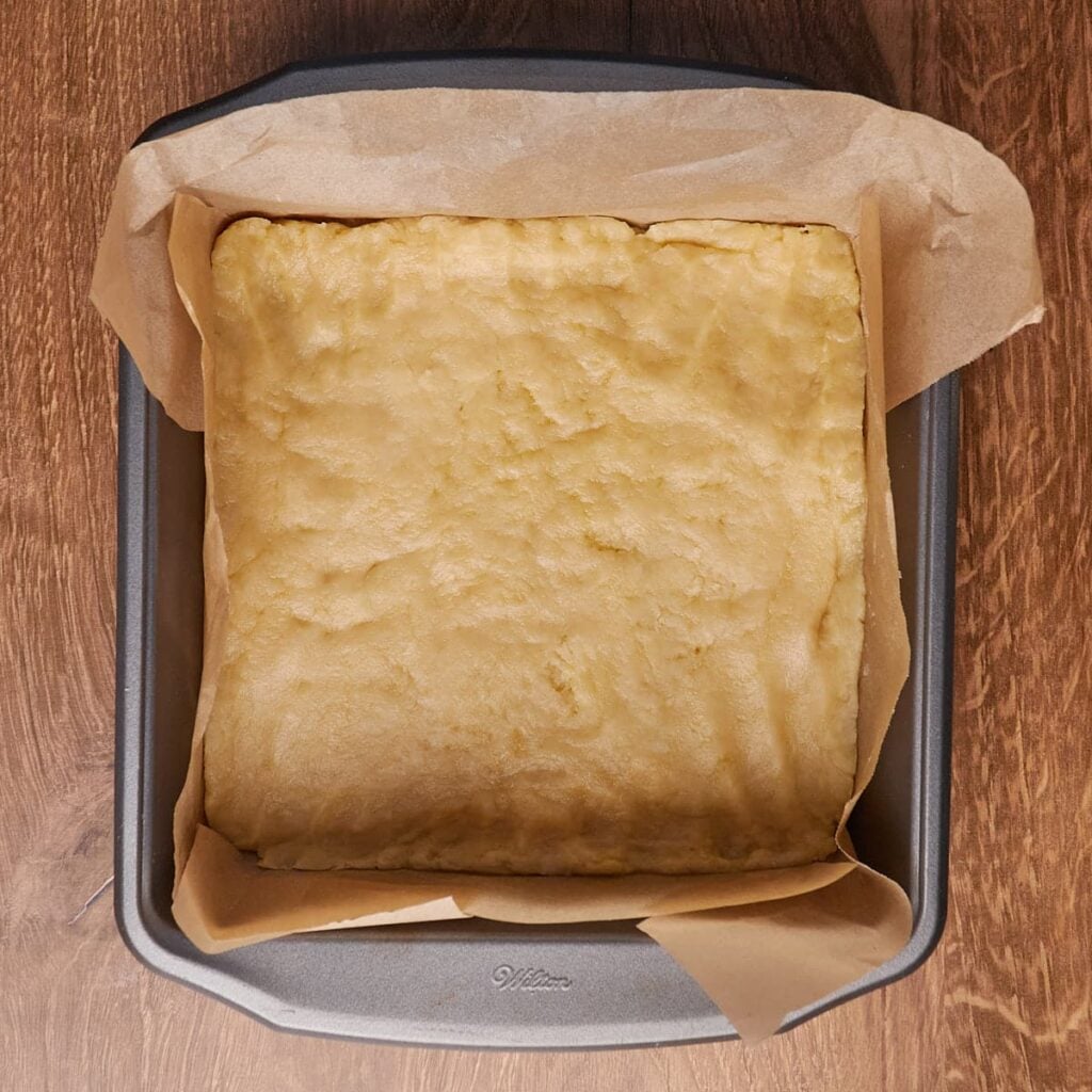 Dough for the shortbread layer of sourdough lemon bars pressed into a 9x9 pan lined with parchment paper.