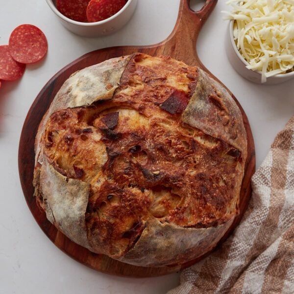 Round loaf of pepperoni and mozzarella sourdough bread on a round wooden board with pepperonis and shredded mozzarella next to it.