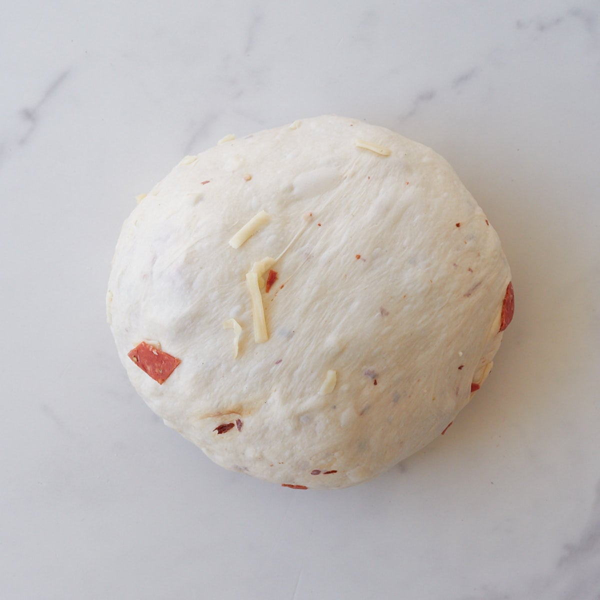 Pepperoni sourdough bread dough in a round ball after being shaped on a white marble counter.