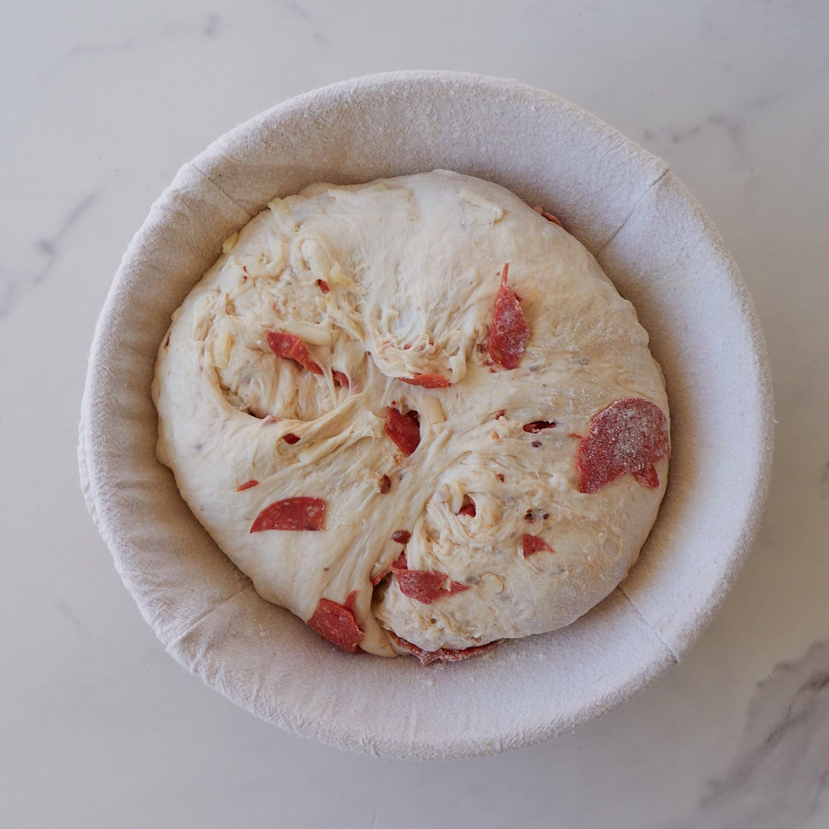 Pepperoni sourdough bread in a round banneton basket on a marble counter.