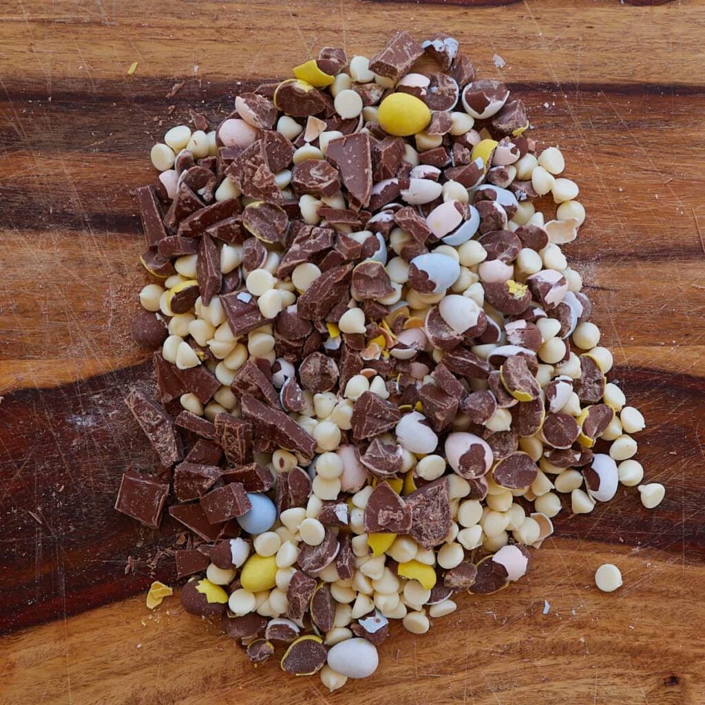 Chopped chocolate, Cadbury eggs and white chocolate chips on a wooden cutting board.
