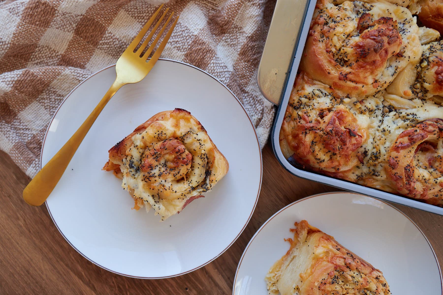 One Sourdough Ham and Cheese Roll on a white plate with a gold fork and a 9x13 pan filled with the rolls next to it