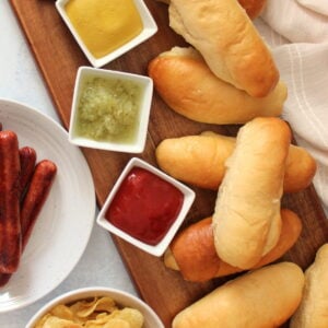 Sourdough discard hot dog buns on a wooden board next to a plate of hot dogs, bowl of chips and small bowls of mustard, relish, and ketchup.