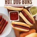 Sourdough discard hot dog buns on a wooden board next to a plate of hot dogs, bowl of chips and small bowls of mustard, relish, and ketchup with three buns with hot dogs inside.