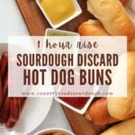 Sourdough discard hot dog buns on a wooden board next to a plate of hot dogs, bowl of chips and small bowls of mustard, relish, and ketchup.
