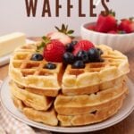 Three sourdough waffles stacked on top of each other on a white plate with blueberries and strawberries on top.