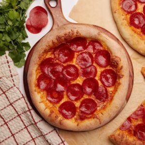 Pepperoni pizza made from sourdough discard pizza dough on a round wooden board next to more pepperoni pizza on parchment paper.