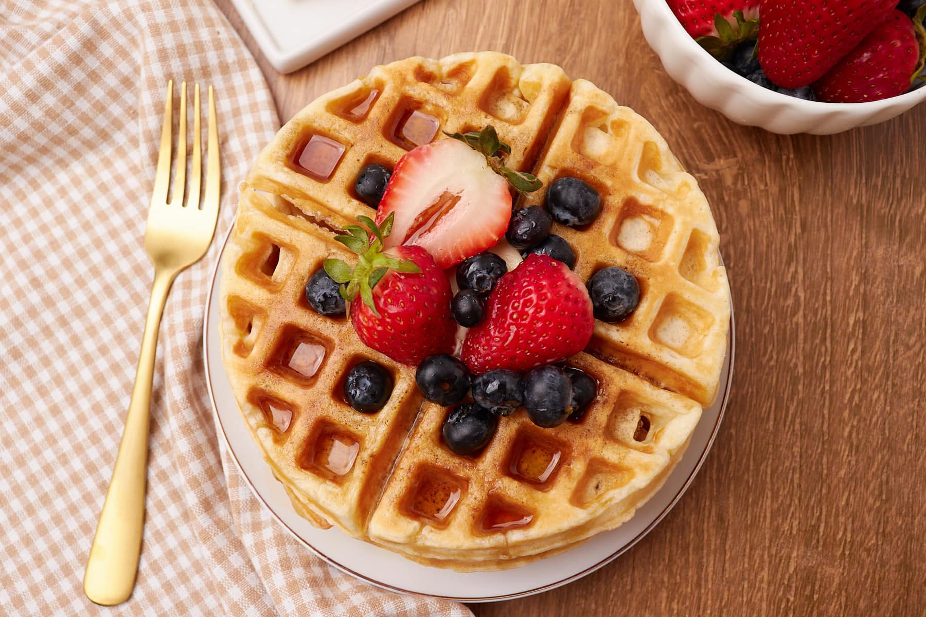 Sourdough Waffles on a white plate with syrup on top and blueberries and sliced strawberries with a gold fork.