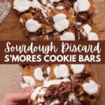 Sourdough S'more Cookie Bars cut into squares on a wooden surface and a hand holding one of the cookie bars.