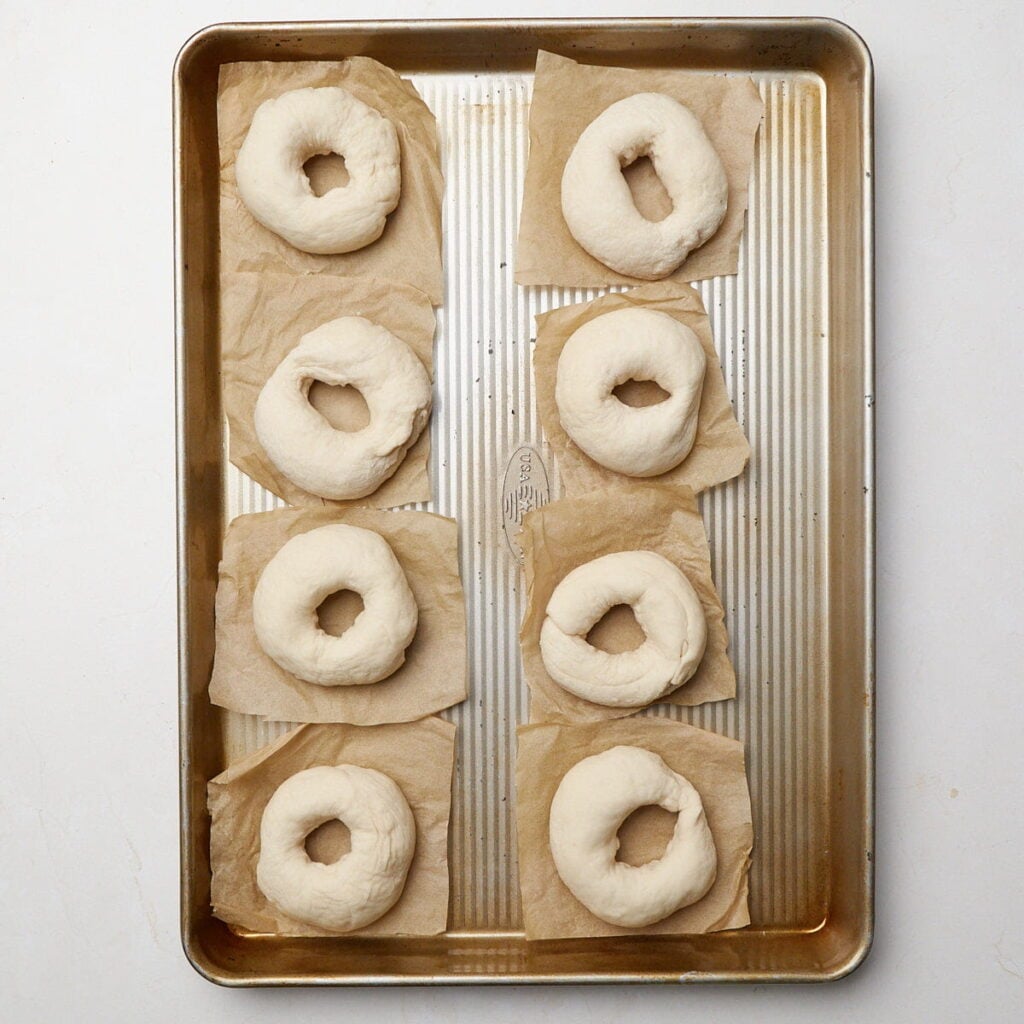 Shaped sourdough discard bagels on a sheet pan on top of parchment paper squares.