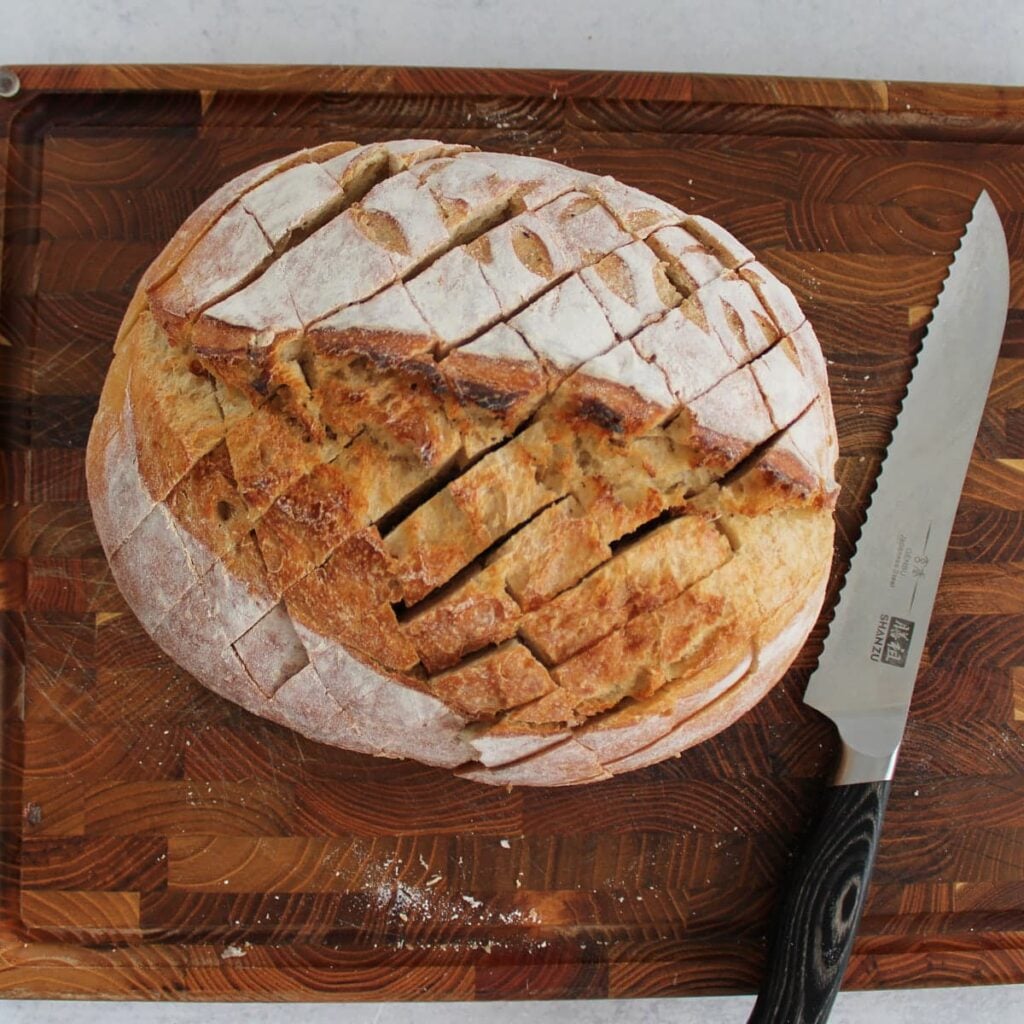 A loaf of sourdough bread on a cutting board next to a bread knife with cuts in it to create the pull apart bread.