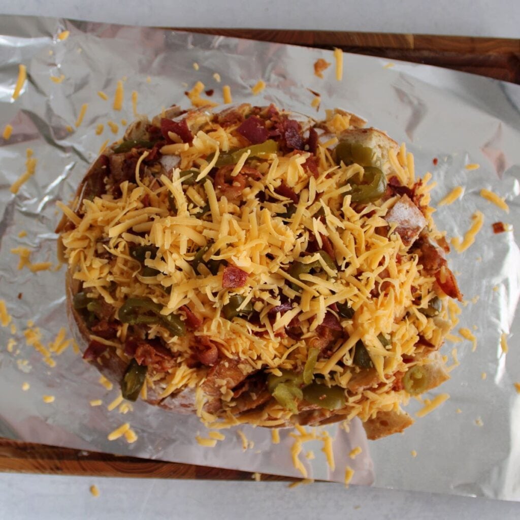 A sourdough loaf sliced and filled with ranch, butter, cheddar cheese, bacon and jalapeños on a piece of foil.