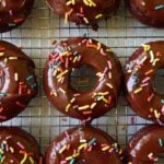 Baked sourdough chocolate donuts with chocolate icing and rainbow sprinkles on a wire rack.