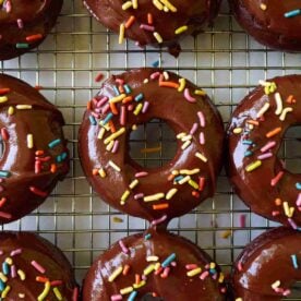 Baked sourdough chocolate donuts with chocolate icing and rainbow sprinkles on a wire rack.