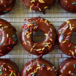 Baked sourdough chocolate donuts with chocolate icing and rainbow sprinkles on a wire rack.