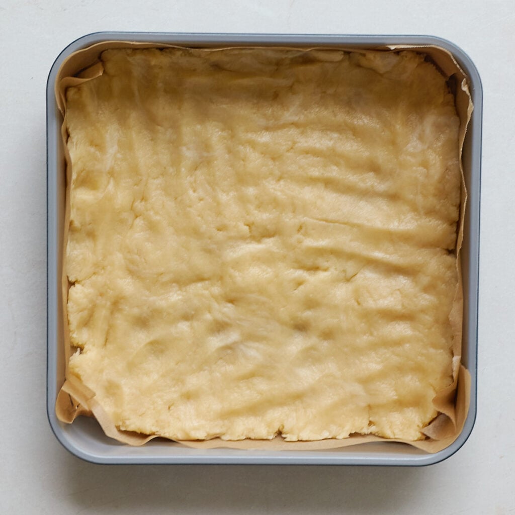 Shortbread layer for Sourdough Peach Crumble Cheesecake Bars pressed into a parchment lined 9x9 baking pan.