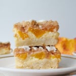 Two Sourdough Peach Crumble Cheesecake Bars stacked on top of each other on a white plate.