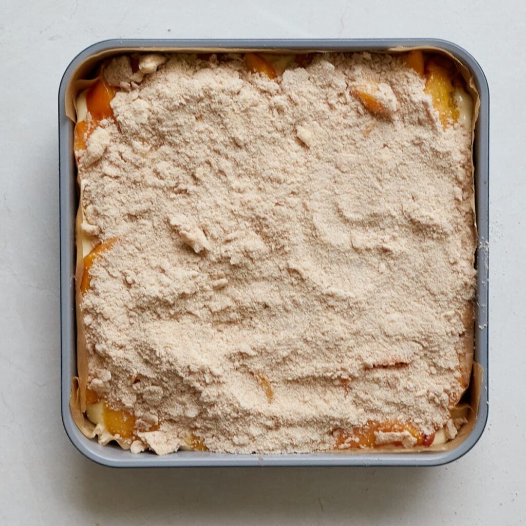Streusel topping added on Sourdough Peach Crumble Cheesecake Bars in a parchment lined 9x9 pan.