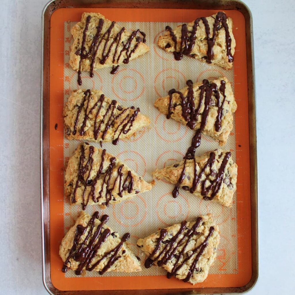 Chocolate drizzle added on top of Chocolate Chip Sourdough Scones on a sheet pan lined with a silicone baking mat.