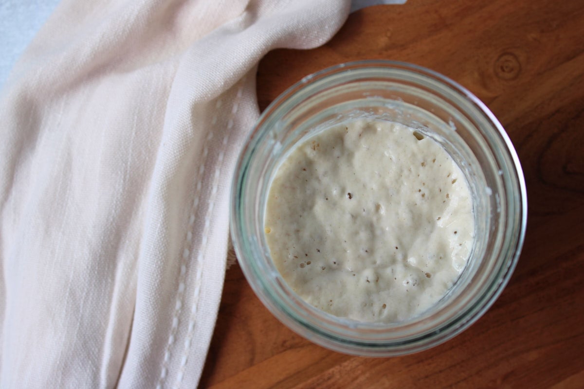 Sourdough starter in a jar with a linen cloth and a wood cutting board.