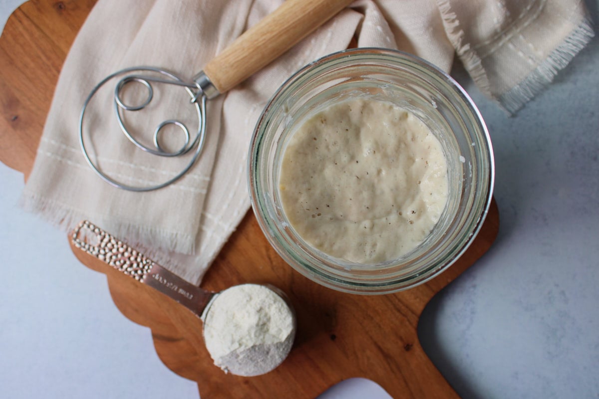 Sourdough starter in a jar next to a measuring cup of flour and a danish dough whisk.