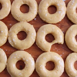 Maple Glazed Sourdough Donuts with flaky salt on top on a wooden board with the middle donut missing a bite.