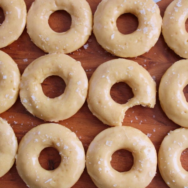 Maple Glazed Sourdough Donuts with flaky salt on top on a wooden board with the middle donut missing a bite.